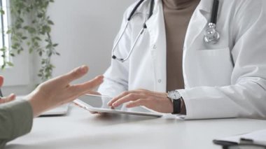 Laboratuvar önlüğü ve steteskop giymiş erkek doktor modern tıp ofisinde tablet kullanırken hastayı dinliyor. İlaç ve sağlık hizmetleri kavramı.