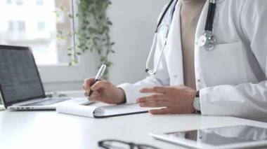 Laboratuvar önlüğü ve steteskop giyen doktor tıbbi reçete yazıyor ya da ofis masasında oturan tıbbi belgeleri dolduruyor. İlaç ve sağlık hizmetleri