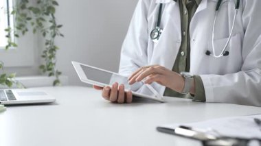 Beyaz laboratuvar önlüğü ve steteskop giyen profesyonel kadın doktor, klinik ortamdaki hasta kayıtlarını incelerken dijital tablet üzerinde çalışıyor. İlaç ve sağlık hizmetleri kavramı
