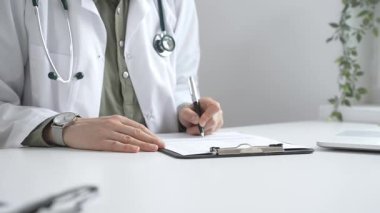 Laboratuvar önlüğü ve steteskop giymiş kadın doktor, tıp bürosunun masasındaki panoya profesyonelliği vurgulayan dizüstü bilgisayar ve gözlüklerle notlar yazıyor. İlaç ve sağlık hizmetleri kavramı