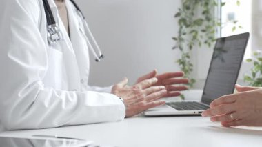 Laboratuvar önlüklü ve steteskoplu doktor, tıbbi ofisindeki dizüstü bilgisayarla hastaya teşhis koymayı açıklarken el kol hareketi yapıyor. İlaç ve sağlık hizmetleri kavramı.