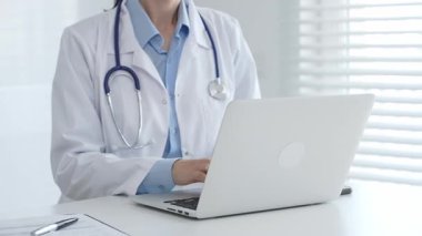 Laboratuvar önlüğü ve steteskop giyen kadın doktor masamda oturmuş bilgisayarında tıbbi belgeler ve kalemle ön planda yazı yazıyor, arka planda panjurları olan parlak bir pencere var.