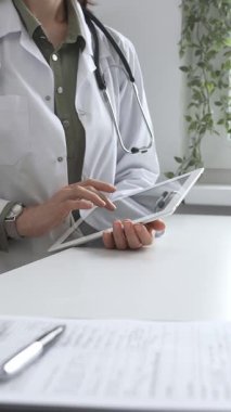 Beyaz laboratuvar önlüğü ve steteskop giyen profesyonel kadın doktor, klinik ortamdaki hasta kayıtlarını incelerken dijital tablet üzerinde çalışıyor. İlaç ve sağlık hizmetleri kavramı