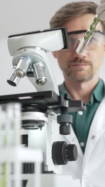 Koruyucu giysiler giyen bilim adamı mikroskop, cam ve bilimsel ekipman yakınında yeşil bitki örnek tüpünü analiz ediyor. Tıp ve bilim konsepti.