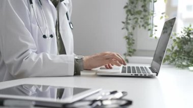 Stetoskop ve laboratuvar önlüğü giyen doktor özenle bir dizüstü bilgisayarda yazıyor ve modern bir tıp ofisinde verimli bir sağlık hizmeti yönetimi sergiliyor. İlaç ve sağlık hizmetleri kavramı