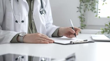 Laboratuvar önlüğü ve steteskop giymiş kadın doktor, tıp bürosunun masasındaki panoya profesyonelliği vurgulayan dizüstü bilgisayar ve gözlüklerle notlar yazıyor. İlaç ve sağlık hizmetleri kavramı