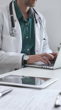 Doktor adam yeşil gömleğin üstüne beyaz önlük giyiyor ve steteskop da parlak bir tıbbi ofisteki dizüstü bilgisayarda yazarak sağlık ve teknolojinin kesişimini gösteriyor. İlaç ve sağlık hizmetleri.