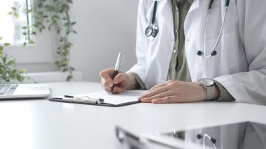 Laboratuvar önlüğü ve steteskop giymiş kadın doktor, tıp bürosunun masasındaki panoya profesyonelliği vurgulayan dizüstü bilgisayar ve gözlüklerle notlar yazıyor. İlaç ve sağlık hizmetleri kavramı