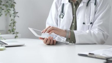 Beyaz laboratuvar önlüğü ve steteskop giyen profesyonel kadın doktor, klinik ortamdaki hasta kayıtlarını incelerken dijital tablet üzerinde çalışıyor. İlaç ve sağlık hizmetleri kavramı