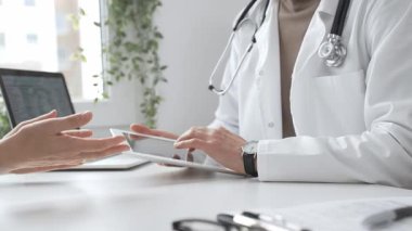 Laboratuvar önlüğü ve steteskop giymiş erkek doktor modern tıp ofisinde tablet kullanırken hastayı dinliyor. İlaç ve sağlık hizmetleri kavramı.