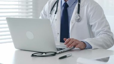 Laboratuvar önlüğü ve steteskop giyen tıp doktoru masasında oturuyor ve güneş gözlüğü ve kalemle yakın bir yerde, parlak bir tıp ofisinde dizüstü bilgisayarda yazıyor. İlaç ve sağlık hizmetleri kavramı