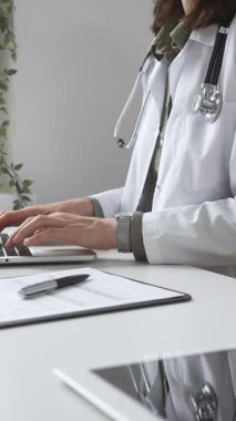 Stetoskop ve laboratuvar önlüğü giyen doktor özenle bir dizüstü bilgisayarda yazıyor ve modern bir tıp ofisinde verimli bir sağlık hizmeti yönetimi sergiliyor. İlaç ve sağlık hizmetleri kavramı