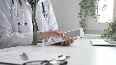 Beyaz laboratuvar önlüğü ve steteskop giyen profesyonel kadın doktor, klinik ortamdaki hasta kayıtlarını incelerken dijital tablet üzerinde çalışıyor. İlaç ve sağlık hizmetleri kavramı