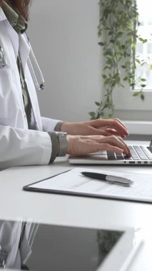 Stetoskop ve laboratuvar önlüğü giyen doktor özenle bir dizüstü bilgisayarda yazıyor ve modern bir tıp ofisinde verimli bir sağlık hizmeti yönetimi sergiliyor. İlaç ve sağlık hizmetleri kavramı