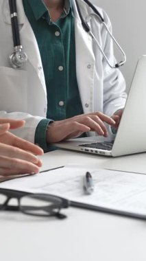 Yeşil gömleğin üzerine laboratuvar önlüğü giyen doktor dizüstü bilgisayarda yazı yazıyor ve klinikteki tıbbi muayene sırasında bir kadının semptomları açıklamasını dinliyor. İlaç ve sağlık hizmetleri kavramı.