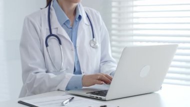 Laboratuvar önlüğü ve steteskop giyen kadın doktor masamda oturmuş bilgisayarında tıbbi belgeler ve kalemle ön planda yazı yazıyor, arka planda panjurları olan parlak bir pencere var.