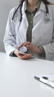 Beyaz laboratuvar önlüğü ve steteskop giyen profesyonel kadın doktor, klinik ortamdaki hasta kayıtlarını incelerken dijital tablet üzerinde çalışıyor. İlaç ve sağlık hizmetleri kavramı