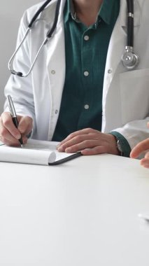 Yeşil gömlekli bir doktor, parlak ve modern bir klinikte muayene sırasında hastanın sağlık sorunlarını dinliyor ve reçete yazıyor. İlaç ve sağlık hizmetleri kavramı