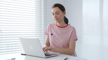 Uzaktan kumandalı profesyonel serbest çalışan, güneş ışığı penceresinin yanındaki dizüstü bilgisayarda kısmen kapalı güneşlik ile yazı yazan, yoğun bir şekilde dijital projeye odaklanan