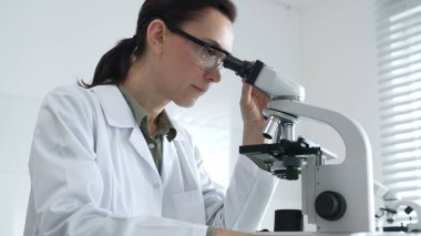 Modern laboratuvar ortamında biyolojik örnekleri incelerken mikroskoba odaklanan koruyucu ekipman takan kadın bilim adamı. Tıp, sağlık ve bilim konsepti