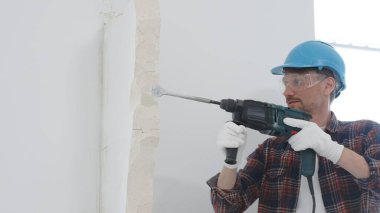 Kırmızı kareli gömlek, mavi başlık ve koruyucu eldiven giyen inşaat işçisi, döner çekiç matkabıyla beyaz duvarı yıkıyor, toz ve portre görüntüsü yaratıyor. Yenileme konsepti.