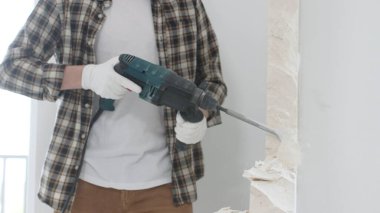 Tanınmayan bej kareli gömlek ve koruyucu eldivenler giyen erkek inşaat işçisi, döner çekiç matkabıyla beyaz duvarı yıkıyor, toz üretiyor, yakın görüş sağlıyor. Yenileme konsepti.