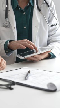 Laboratuvar önlüklü doktor yeşil gömlekli ve steteskop modern tıp bürosunda hastaya danışmanlık yaparken tablet kullanıyor. İlaç ve sağlık hizmetleri kavramı.