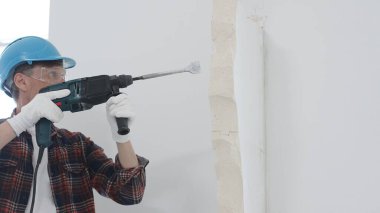Kırmızı kareli gömlek, mavi başlık ve koruyucu eldiven giyen inşaat işçisi, döner çekiç matkabıyla beyaz duvarı yıkıyor, toz ve portre görüntüsü yaratıyor. Yenileme konsepti.
