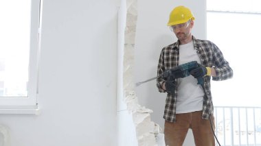 Bej kareli gömlek, sarı başlık ve koruyucu eldiven giyen inşaat işçisi, dönen çekiç matkabıyla beyaz duvarı yıkıyor ve orta uzunlukta toz üretiyor. Yenileme konsepti