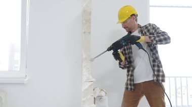 Bej kareli gömlek, sarı başlık ve koruyucu eldiven giyen inşaat işçisi, dönen çekiç matkabıyla beyaz duvarı yıkıyor ve orta uzunlukta toz üretiyor. Yenileme konsepti