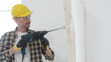 Bej kareli gömlek, sarı başlık ve koruyucu eldiven giyen bir inşaat işçisinin portresi, döner çekiç matkabıyla beyaz duvarı yıkıyor ve toz oluşturuyor. Yenileme konsepti.