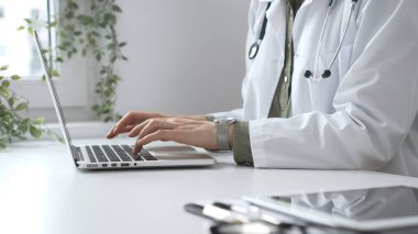 Stetoskop ve laboratuvar önlüğü giyen doktor özenle bir dizüstü bilgisayarda yazıyor ve modern bir tıp ofisinde verimli bir sağlık hizmeti yönetimi sergiliyor. İlaç ve sağlık hizmetleri kavramı