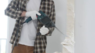 Tanınmayan bej kareli gömlek ve koruyucu eldivenler giyen erkek inşaat işçisinin yakın çekiç matkabıyla beyaz duvarı yıkıyor, toz üretiyor, yakın görüş sağlıyor. Yenileme.