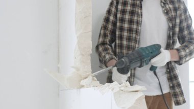 Tanınmayan bej kareli gömlek ve koruyucu eldivenler giyen erkek inşaat işçisi, döner çekiç matkabıyla beyaz duvarı yıkıyor, toz üretiyor, yakın görüş sağlıyor. Yenileme konsepti.