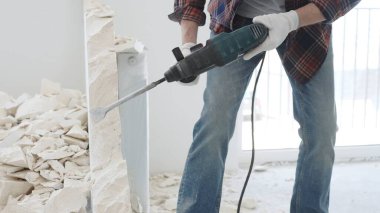 Kırmızı kareli kotlar ve koruyucu eldivenler giyen kimliği belirsiz erkek inşaat işçisi, döner çekiç matkabıyla beyaz duvarı yıkıyor, yerde enkaz oluşturuyor, manzarayı kapatıyor. Yenileme.