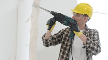 Bej kareli gömlek, sarı başlık ve koruyucu eldiven giyen inşaat işçisi, döner çekiç matkabıyla beyaz duvarı yıkıyor, toz ve portre görüntüsü yaratıyor. Yenileme konsepti.