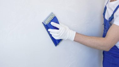 Koruyucu beyaz eldivenler ve mavi inşaat tulumları giyen kimliği belirsiz erkek inşaat işçisi, iç dekorasyon sırasında pürüzsüz yüzey hazırlamak üzere mavi el bloğuyla beyaz duvarı zımparalıyor.