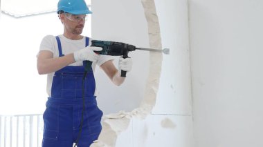 Mavi tulumlu ve koruyucu beyaz eldivenli bir inşaat işçisi, beyaz duvarı döner çekiç matkabıyla yıkıyor, toz üretiyor, yatay görüşü kapatıyor. Yenileme konsepti.