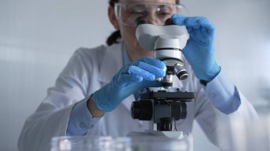 Laboratuvar önlüğü, beyaz koruyucu eldiven ve gözlük giyen kadın bilim adamı modern laboratuvar düzeninde mikroskop kullanıyor, portre görüntüsü. Gösterişsiz ışıklandırma fotoğrafı. Bilim ve Tıp.