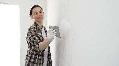 Koruyucu eldiven giyen ve kareli gömlekli bir kadın beyaz duvara macun yapıştırıyor, ev tadilatı sırasında spatulayla bileşik yayıyor, pratik ev geliştirme becerileri sergiliyor..