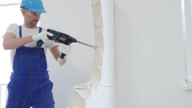 Mavi tulumlu, koruyucu şapkalı ve beyaz eldivenli bir inşaat işçisinin portresi, döner çekiç matkabıyla beyaz duvarı yıkıyor, toz oluşturuyor, yatay görüş sağlıyor. Yenileme.
