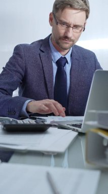 Mavi takım elbiseli iş adamı ofis setindeki cam masada dizüstü bilgisayar, hesap makinesi ve belgelerle çalışıyor. Düşük ışıklandırma, dikey portre. İş adamları ve denetim.