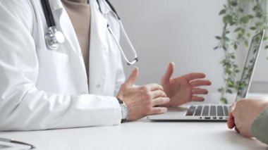 Laboratuvar önlüklü ve steteskoplu doktor, tıbbi ofisindeki dizüstü bilgisayarla hastaya teşhis koymayı açıklarken el kol hareketi yapıyor. İlaç ve sağlık hizmetleri kavramı.
