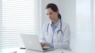 Laboratuvar önlüğü ve steteskop giyen kendine güvenen kadın doktor, doğal ışık dolu, parlak ve modern bir muayenehanede çalışırken laptopuna odaklanmış bir şekilde bir masada oturuyor.