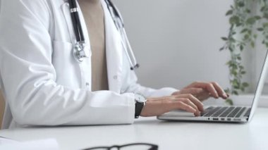 Laptop üzerinde çalışan, modern, iyi aydınlatılmış sağlık ortamında beyaz önlük ve steteskop giyen, klinik uzmanlığı dijital teknolojiyle harmanlayan bir tıp uzmanı. İlaç ve sağlık hizmetleri