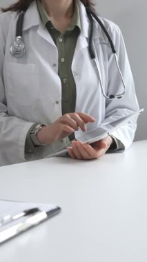 Beyaz laboratuvar önlüğü ve steteskop giyen profesyonel kadın doktor, klinik ortamdaki hasta kayıtlarını incelerken dijital tablet üzerinde çalışıyor. İlaç ve sağlık hizmetleri kavramı
