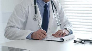 Laboratuvar önlüğü ve steteskop giyen erkek doktor, tıbbi bir ofiste bir masada oturuyor, hasta bakımı ve tedavisi için bir not panosuna not alırken dizüstü bilgisayar kullanmaya odaklanıyor.