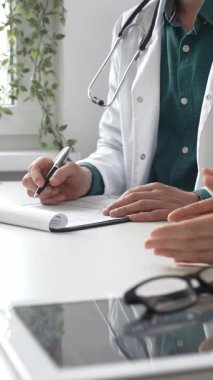 Yeşil gömlekli bir doktor, parlak ve modern bir klinikte muayene sırasında hastanın sağlık sorunlarını dinliyor ve reçete yazıyor. İlaç ve sağlık hizmetleri kavramı