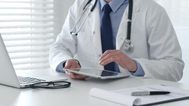 Dijital tablet üzerinde çalışan, beyaz laboratuvar önlüğü ve steteskop giyen, dizüstü bilgisayarı, gözlüğü ve hasta tablosu olan bir tıbbi uzman. İlaç ve sağlık hizmetleri kavramı