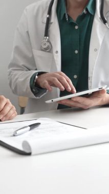 Yeşil gömlekli bir erkek doktor ve modern tıp ofisinde tablet kullanan hastaya teşhisi açıklarken steteskop hareketi yapıyor. İlaç ve sağlık hizmetleri kavramı.
